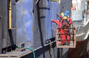 Ship crew performing hull maintenance on a vessel, using safety harnesses and scaffolding during ship husbandry operations in port.